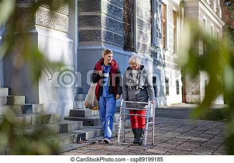 Senior Woman With Walking Frame And Caregiver Outdoors In Town