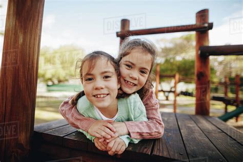 Little girl hugging her twin sister while lying in a wooden structure at playground. Beautiful ...