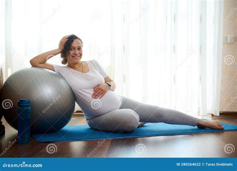 Cute Pregnant Woman Sitting On Exercise Mat Strokes Her Belly Having Rest In Fitness Workout
