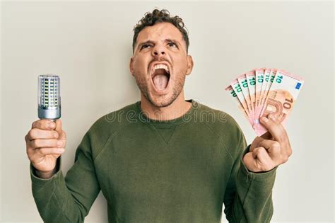 Young Hispanic Man Holding Led Bulb And Euros Banknotes Angry And Mad