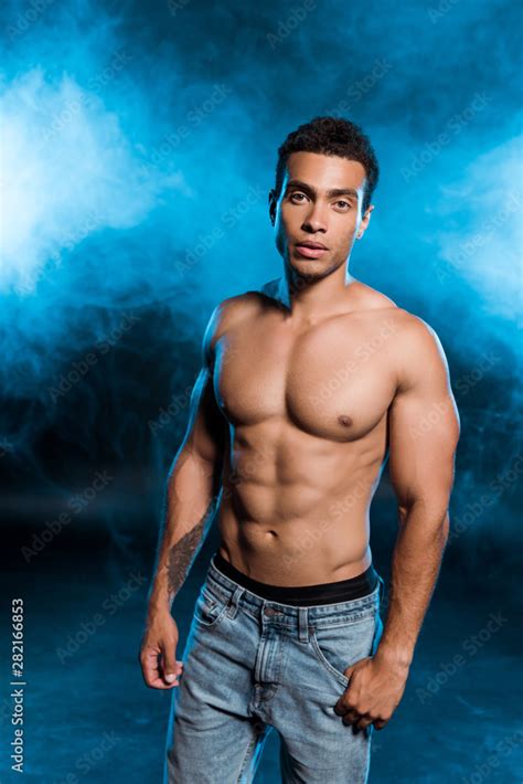 Sexy Mixed Race Man Standing In Denim Jeans And Looking At Camera On Blue With Smoke Stock Photo