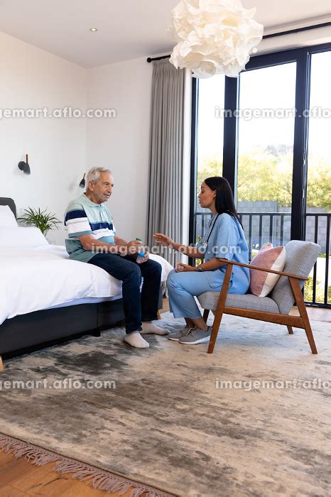 Biracial Female Physiotherapist Talking With Caucasian Senior Man Holding Medicines On Bed At
