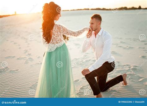 Groom Stands Barefoot Before Bride On One Knee In Desert On Suns Stock