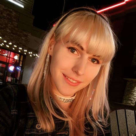 A Woman With Blonde Hair Wearing A Black Jacket And Gold Earrings Smiling At The Camera