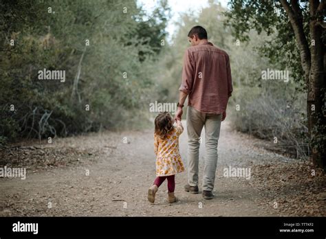 Rear View Of Father And Daughter Holding Hands While Walking On Dirt