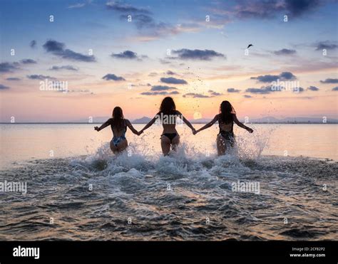 Back View Of Playful Slim Girlfriends Running In Water In Bikini At Sunset In Summer And Holding