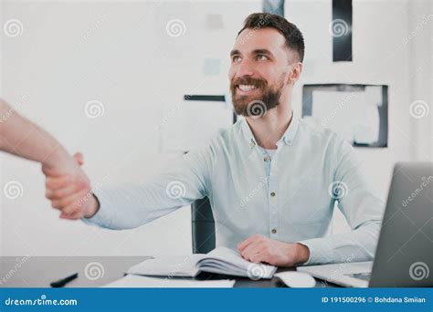 Young Bearded Smiling Man In Elegant Shirt Sits In Modern Bright Office Shaking Hands With