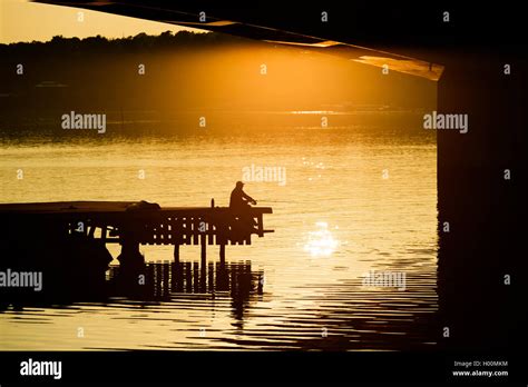 Silhouette Of Man Sitting On Pier In The Evening Sunlight While Fishing